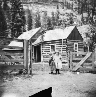 Two small girls stand in the open gateway of a wood fence, in front of a log house, Ouray, Ouray County, Colorado.