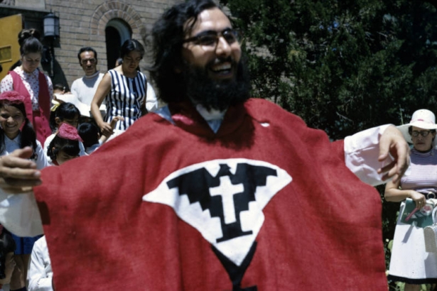 Father José Lara stands outside Our Lady of Guadalupe Church, 1209 West 36th Avenue, in the Highland neighborhood, Denver, Colorado. He holds his arms outstretched to display the UFW Aztec eagle on his vestments. Young men and women leave the church in the distance.
