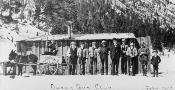 The Ouray Gun Club poses with their rifles outside a board-and- batten shack on a snowy day north of Ouray, Ouray County, Colorado. They include, left to right: Gus Arps in cart drawn by a small burro, unidentified man, Albert Arps, Otto Arps, Charley Pearson, Barney Du Praw, Andy Sneva, Ed Arps, Alf Armstrong, George Armstrong (little boy.)