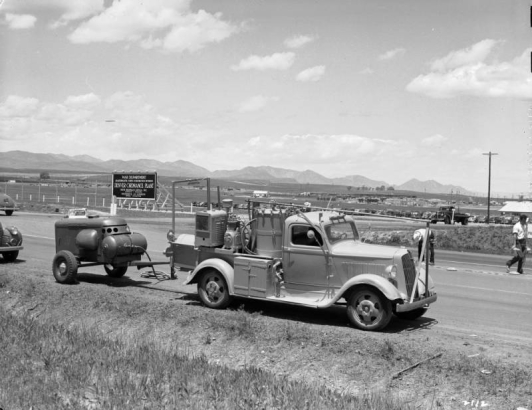 View of the Remington Ordnance Plant (later called the Federal Center) in Lakewood (Jefferson County) Colorado; men install road reflectors by a (probably) striping truck, with a compressor in tow. Sign reads: "War Department - Quartermaster Corps - Construction Division - Denver Ordnance Plant - Smith Henchman & Grylls, Inc., Broderick and Gordon General Contractors." Front range mountains and clouds are in the distance.