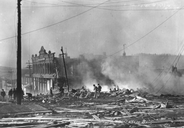 Destruction on Bennett Avenue caused by second fire of April 29, 1896, Cripple Creek, Colorado; groups of men poke through smouldering remains and debris; scene includes burned-out two-story Tutt and Penrose building with flat roof and ornamental parapet.