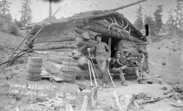 Three men pose by a log cabin belonging to S. J. Roberts, Cripple Creek, Colorado; one man with pants tucked into knee-high boots stands near miner's pick, broom, and sledge hammer (possibly S. J. Roberts); men sit holding a flute or fife and a fiddle or violin; a rack of elk's antlers is over the door.