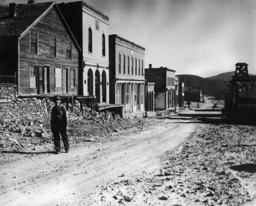 Leonard Nichols grins and walks on Main Street in Nevadaville, Gilpin County, Colorado. A residence tops a stone retaining wall behind him. The Masonic Lodge has a brick front with arched windows on both floors and an incised panel under the cornice. The Oddfellows Building has stone trim and storefronts with millworked pilasters. The Bon-Ton Saloon has three arches. The wood-frame tower of the City Hall/Fire Department has a bell; cars are parked in the background.