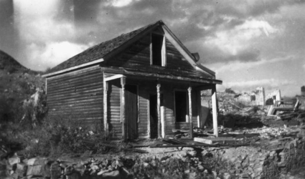 The Simmons house, on Letcher Street, in Nevadaville, Gilpin County, Colorado, is a dilapidated one story frame structure with a gable roof, a covered porch, and lathe-turned piers. Door and window openings are  empty or covered by corrugated tin; debris covers the ground, and ruins of  stone walls are in the background.