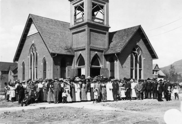 The congregation of Paonia Methodist Church stands outside of the church in Paonia, Colorado in Delta County.  Architectural features of the L-shaped, masonry building: gable roof, gothic windows, fish-scale siding, and a belltower with a hipped roof.
