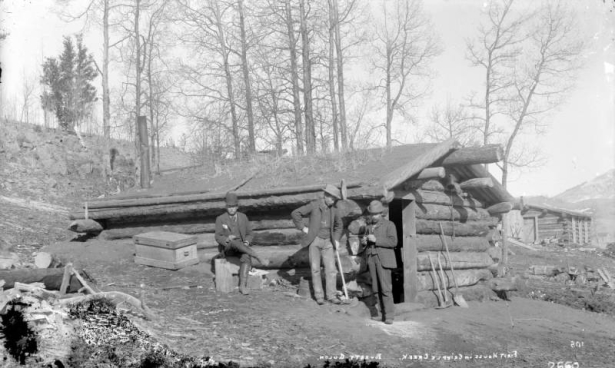 First house in Poverty Gulch, Cripple Creek, Colorado; house is possibly Robert Womack's cabin in Fremont (prior to Cripple Creek incorporation) or Andy Frazier's cabin in Squaw Gulch; the one-story rounded log cabin built into the hillside has a low gable sod roof;  three men wearing three-piece suits and knee high boots pose outside cabin; one man leans on miner's pick while another man, wearing a derby hat, sits on stump; additional picks and shovel are near front side entrance of cabin.