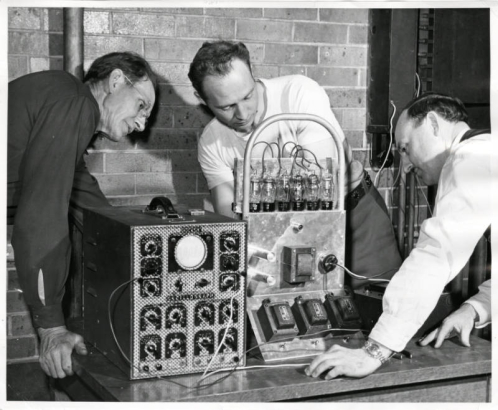Photograph of three men during a radio repair class at the Emily Griffith Opportunity School located in Denver, Colorado.  Two men watch while a man in the center stands behind the machine working on it.