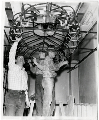 Photograph of three unidentified men during a class in ceiling construction at the Emily Griffith Opportunity School located in Denver, Colorado.  The three men stand on platforms and are working on the curved oval structure.