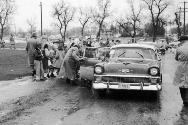 Children stand on a sidewalk beside cars after an evacuation at Lakewood Elementary School in Lakewood (Jefferson County), Colorado. Children and teachers or parents wear rain coats and scarves or hats. A license plate reads: "D11 3818, Colorado, '57."