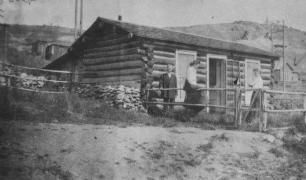 Log cabin residence of Martha and Mary Crane, Cripple Creek, Colorado; one woman sits on upper part of split rail fence, another woman stands near fence gate and one man stands in front of cabin; former residence of Ralph Carr, ex-Governor of Colorado is in the background; residence includes stone fence or foundation, low gable roof with shingles,  single entry at center, opened screen and front door, two double-sash plain windows; background includes outbuildings, mine tailings and surface buildings.