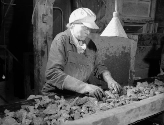 A woman worker sorts through rocks on a conveyor belt at a mill building for a London Mine near Alma (Parker County), Colorado. She wears work overalls, heavy gloves, and a cap. Glasses rest on her nose.