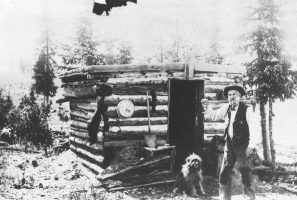 "Old Man Topping" with a miner's pick over his shoulder and his dog pose in front of first log cabin in Cripple Creek, Colorado; the cabin is small, flat-roof and rounded with a gold pan on exterior cabin wall and a shovel propped against cabin wall.