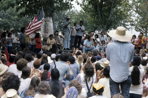 United Farm Workers of America members and supporters hold a rally for the lettuce workers who are on strike against the Finerman Company in Center (Saguache County), Colorado. Men, an African American and a Hispanic American, stand on a stage and men and women look on. Someone holds an American flag and a banner of Our Lady of Guadalupe is tied to a tree.