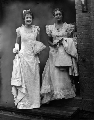 Women pose in a Teller House doorway, in Central City, Gilpin County, Colorado. They wear long, sleeveless wedding dresses; one has a smocked organdy blouse, a sheer overskirt, flounces, and ruffled shoulders.  The other has a brocade front, a square collar, lace shoulders, a train, and a glove. Both hold fans; one wears a cameo and a baguette.
