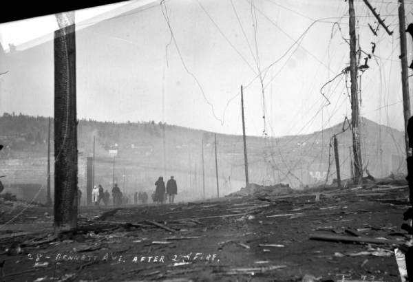 View of Bennett Avenue's desolation and total destruction caused by second fire of 1896, Cripple Creek, Colorado; destruction includes burnt telegraph or utility poles and scattered debris of timbers; groups of men survey remains and destruction; Mount Pisgah is on distant horizon.