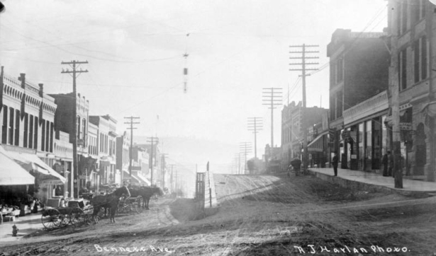 Street and terrace view of one and two-story flat-roof brick commercial buildings on Bennett Avenue, Cripple Creek, Colorado; row of horse-drawn wagons are parked alongside sidewalk; scene includes fire hydrant, awnings, numerous signs attached to buildings, row of utility poles on both sides of street, and wooden stairway with metal railing alongside terrace.