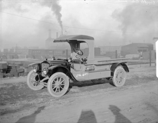 View of a truck used by the Mountain State Telephone and Telegraph Company in Pueblo (Pueblo County), Colorado. The truck has a hand brake, sirens, and plastic windshield rolled and attached to the roof of the automobile. Words painted on the side of the truck read: "Mountain States Tel. & Tel. Co." Smoke emerges from the smokestack of the Colorado Fuel and Iron Company steel mill in the distance.