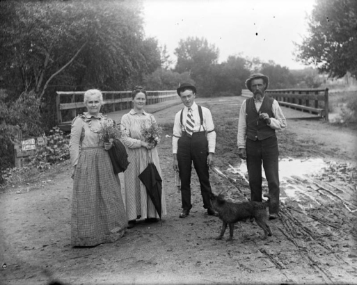Outdoor portrait of two couples on Alameda Avenue in Denver, Colorado. The women hold bouquets of flowers. One man wears suspenders and an armband. A dog and the Alameda Avenue bridge over the South Platte River are nearby.
