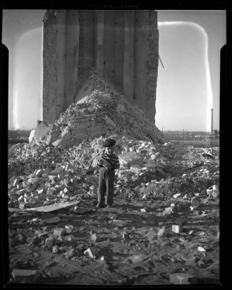 Photograph of a small boy standing in the debris of the Omaha-Grant smoke stack after its demolition. The boy stands at the base of the ruins, carrying a brick away from a pile of debris.