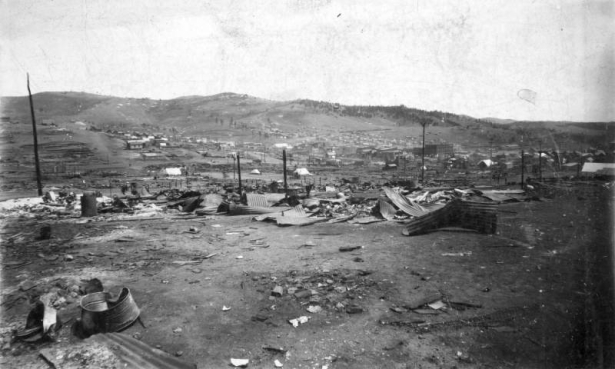 View of destruction caused by 1896 fire, Cripple Creek, Colorado; scene includes remains of twisted sheet metal, ashes and burnt timbers, and former business and commercial district; canvas tents are scattered amidst fire debris; remaining buildings and residences are in the distance on the hillside.