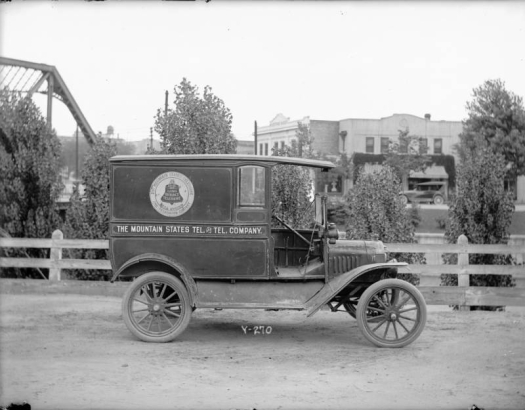 View of a Ford delivery truck used by the Mountain States Telephone and Telegraph Company in Denver, Colorado. A bell logo is painted on the side of the truck and lettering reads: "Mountain States Tel. & Tel. Company, Local and Long Distance Telephone, Bell System." Businesses and bridges are in the distance.