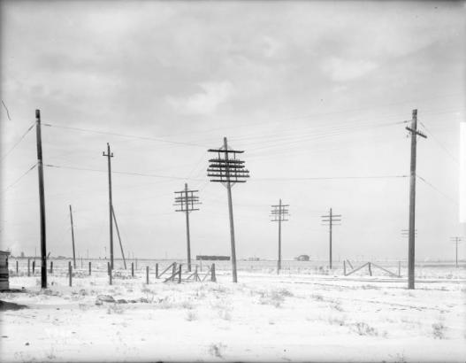 View of telephone poles along a dirt road in Frederick (Weld County), Colorado. The poles have multiple cross arms. Shows barbed wire fences and open fields.