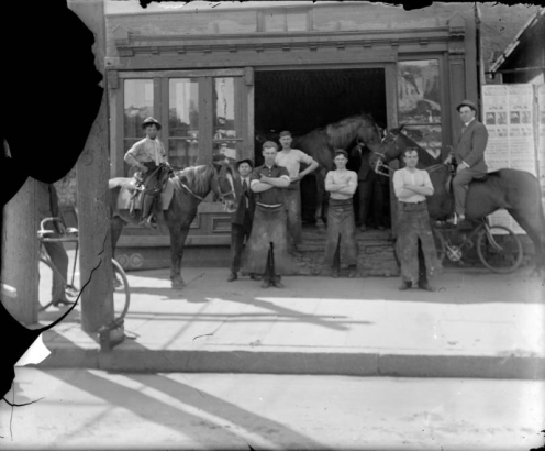 A group of men pose near possibly a blacksmith's shop in Denver, Colorado. Two men are on horseback and other men wear long aprons.