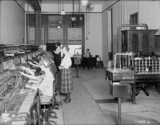 Interior view of the Mountain States Telephone and Telegraph Company exchange in Denver, Colorado; shows women working. A woman in a plaid dress and low heeled shoes is by signs: "Miss Fitzsimmons," and "Miss Andrews."