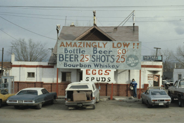 A woman poses in front of Dan's Corner Tavern,  5302 Brighton Boulevard in the Elyria-Swansea neighborhood of Denver, Colorado. The bar, a former residence, has a cross-gabled roof, brick facing and several additions. Signs read, "AMAZINGLY LOW!!, Bottle Beer 60 cents, BEER 20 cents, SHOTS 25 cents, Bourbon Whiskey, Pete Staffieri," "EGGS, SPUDS," "NOTARY PETE" and "Notary Public, Bottle Beer." A phone booth is near the building and cars are parked nearby.
