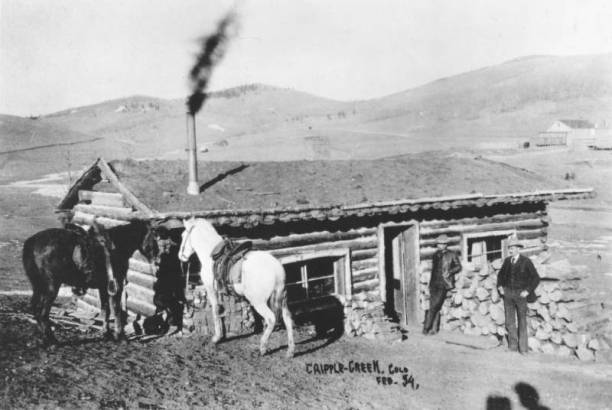 Harry Leonard and Spencer Penrose stand in front of Tutt and Penrose cabin, Cripple Creek, Colorado; one-story log cabin built in 1891 with low gable and sod roof; two saddled horses are teethered to corner of cabin; cabin includes stovepipe and wooden plank leading to front door; sparsely settled hillsides are in the background.