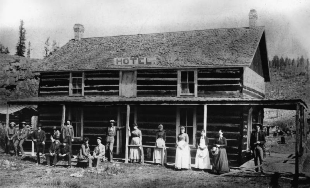 Women, men, and A. B. Cooper (miner), pose in front of a hotel in Whitepine, a silver camp in Gunnison County, Colorado. The hewn log building has a covered porch and sign: "Hotel."