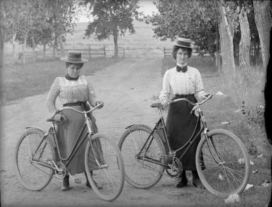 Women pose with their bicycles in a park possibly in Denver, Colorado. They wear dresses and straw wide-brimmed hats.