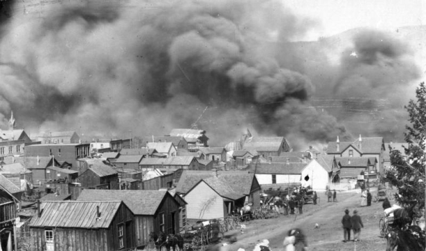 View of April 29th, 1896, fire and clouds of smoke from junction of Myers Avenue and "A" Street after explosion at El Paso Livery stable, Cripple Creek, Colorado; scene includes horse-drawn wagons with household goods or personal belongings parked in front of small wooden frame residences (probably cribs of infamous red-light district of Myers Avenue); men and women are in front of residences and on the dirt street.