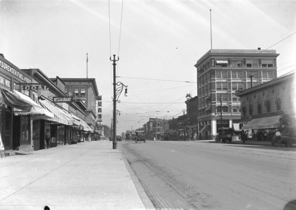 View of Broadway at Ellsworth Street, looking north in Denver, Colorado. Multi-story  commercial buildings, street lamps and street railway tracks line the street. Notable buildings include the Broadway Bank building and the Queen Theater. A person rides a bike and people stand near storefront windows and under awnings.  Automobiles are parked nearby. Signs on buildings read: "Restaurant," "William Small, Groceries & Meats," "Coal," "Sorensen Fuel and Feed" and "Queen."