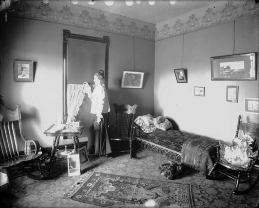 Interior view of a bedroom, probably in Colorado. A woman, in a dress, holds a book and stares out the window. Items in the room include a bed with pillows and a blanket, rocking chairs, an ornate rug, patterned carpet, wall furnishings and a table with framed portraits.