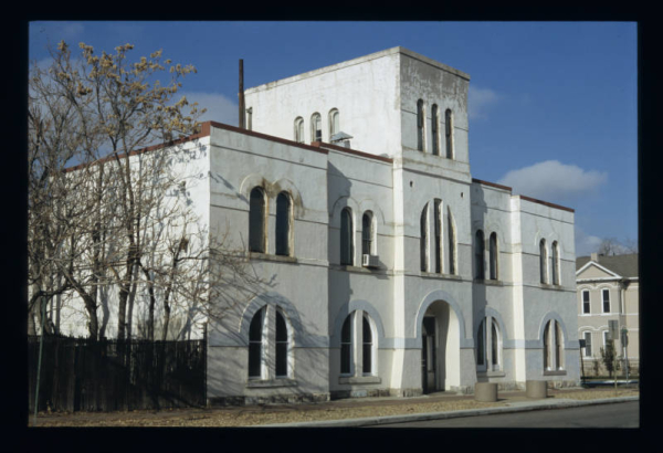 View of the State Armory (1889; William Quayle) at 2565 Curtis Street in the Five Points neighborhood of Denver, Colorado. Shows a two-story, brick, Romanesque Revival influenced, symmetrical building with semi-circular arched windows, belt courses and an inset entrance in a three-story tower. The brick walls have been stuccoed. The former armory is part of the Curtis Park-Champa Street Historic District and now houses Scientific Site Inc.