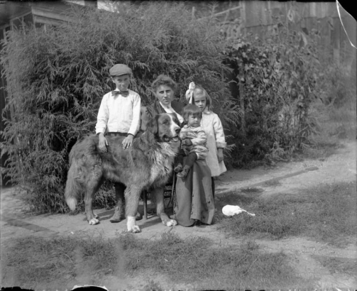 Outdoor portrait of a woman, her sons, daughter, and dog in a garden in Denver, Colorado. The girl wears a ribbon in her hair and one son wears a bow tie and cap.