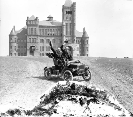 Harry M. Rhoads, behind the steering wheel, poses in a convertible with passenger in front of Westminster University or College, at 3455 West 83rd Avenue in Westminster (Adams County), Colorado. Animals graze in the barren field in which the building is situated. Architectural features of the building: sandstone construction, 175-foot tower with a hipped roof, conical towers, square towers, arched and square windows and entrances, gables, and chimneys.