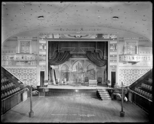 Interior view of El Jebel Temple, Denver, Colorado; local Masonic order of El Jebel Shriners (building dedicated November, 1907); shows stage area of auditorium with chairs and bench on stage, backdrop, curtain draped open, wall mural painted by Jens Eriksen, ceiling with light bulbs arranged to depict stars, upright piano, radiators, and potted palm trees. The temple was designed by the Baerresen Brothers.