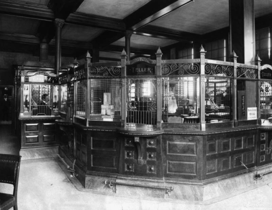 Interior view of the First National Bank, Colorado Springs, El Paso County, Colorado, features coffered wood ceilings and an ornate, three-windowed tellers box and brass footrails under writing shelves. A. H.  Hunt, the cashier is behind beaded grating and another man stands in the background. Signs read: "Cashier," "Teller," "Next Window," and "Revenue Stamps for sale at This window."