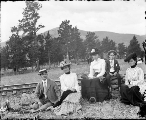 Group portrait of a man, women, and a boy at a picnic in Colorado; costume includes hats with flowers. Hills and railroad tracks are in the background.