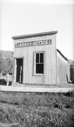View of the dilapidated Assay Office in Pitkin, Gunnison County, Colorado, a wooden, false front building with vertical board siding and a plank sidewalk surrounded by unkempt weeds and grass.