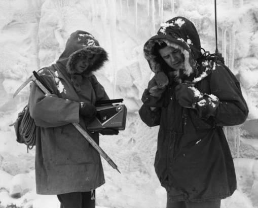 Two AdAmAn club members stand in front of a ice wall with icycles and prepare for the annual New Year's Eve Pikes Peak climb, Colorado Springs, El Paso County, Colorado. The men wear parkas and mittens; one has a backpack with an antenna, the other holds an ice- climbing pick, a "Motorola," portable radio and a coiled rope.