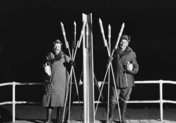 Members Garrett and MacDonald of the AdAmAn Club pose with fireworks including rockets and bombs with fuses, on top of Pikes Peak, Colorado Springs, El Paso County, Colorado. They wear tall lace-up boots and fur collared parkas.