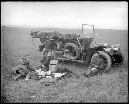 Three men picnic next to an early gasoline-powered automobile near dam site for Standley Lake, Colorado; men eat apples and sit on car seat cushions; scene includes blanket, wicker picnic basket, food items, plates, bottles, a spare tire attached to automobile runner, and the phographer's box of supplies in backseat. McClure photographed construction of Standley Lake by Kenefick-Quigley-Russel Construction Company.