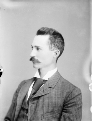Studio bust portrait of a man with a moustache and short hair brushed straight up, in a plaid suit, a silk tie and a watch chain.