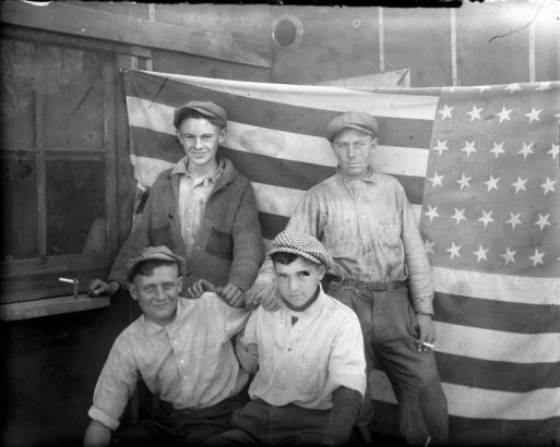 Group portrait of teenaged boys by a United States flag in Denver, Colorado; costume includes duster hats, a knit sweater, and a cigarette.