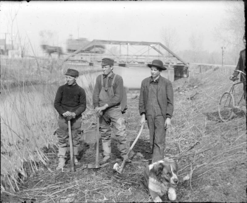 Teenaged boys with axes pose by Archer Canal in Denver, Colorado. A dog and a man with a bicycle are nearby. A horse-drawn wagon makes its way over the Alameda Avenue bridge in the distance.