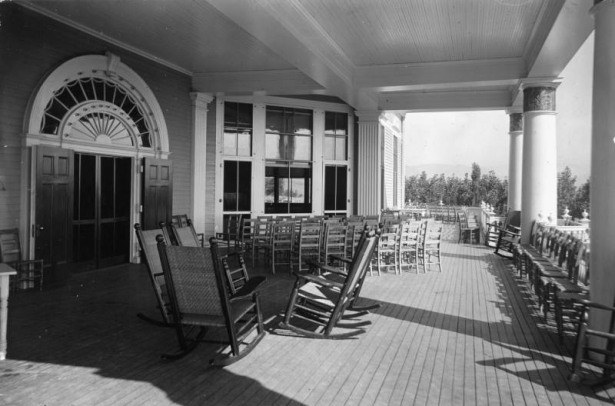 Deck and rocking chairs are arranged on the balcony of the Casino at the Broadmoor Hotel and Casino, Colorado Springs, El Paso County, Colorado. A fanlight tops double doors; columns with ornate capitals support the beamed ceiling.