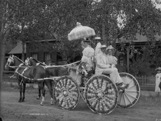 Business partners Charles L. Tutt and Spencer Penrose, Mrs. Tutt,  and Nina Crosby pose in a flower decorated buggy for the Sunflower Carnival Parade, Colorado Springs, El Paso County, Colorado. The men wear white suits and straw hats; the women wear flowered hats and carry a ruffled parasol.